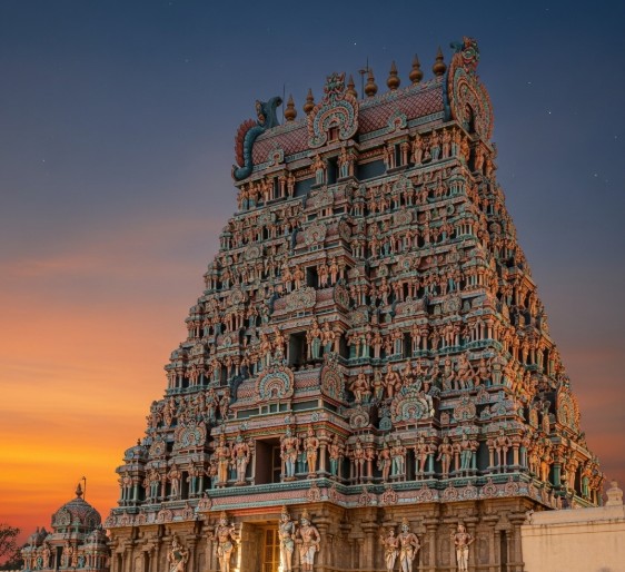 An elaborate, towering gopuram of the Meenakshi Amman Temple is shown at twilight. The structure is covered with thousands of colorful, intricately carved mythological figures. The sky behind it is a blend of deep orange and dark blue, with a few stars visible.