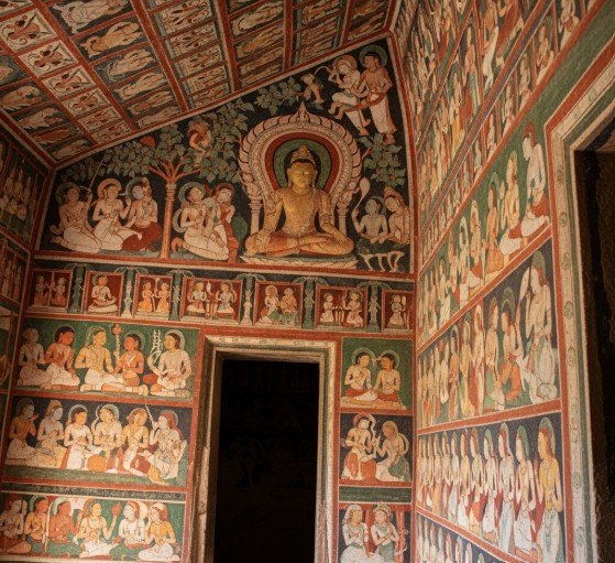 A view of a corner inside an Ajanta Cave, where the walls and ceiling are covered in ancient murals. The colorful paintings, depicting various scenes and figures, are arranged in a grid-like pattern. A central, larger mural shows a seated Buddha figure. The artwork appears to be illuminated by soft, natural light from a doorway.